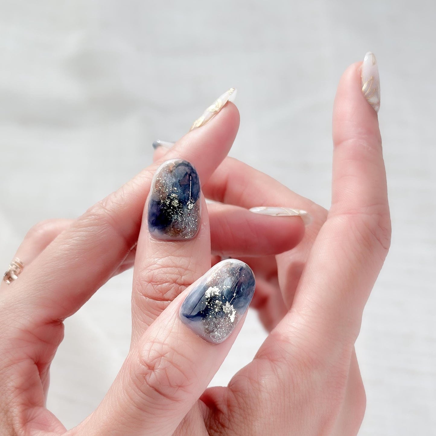 Hand with Oval-shaped press-on nails with deep navy and gold marble art, gold foil accents, and soft floral details.
