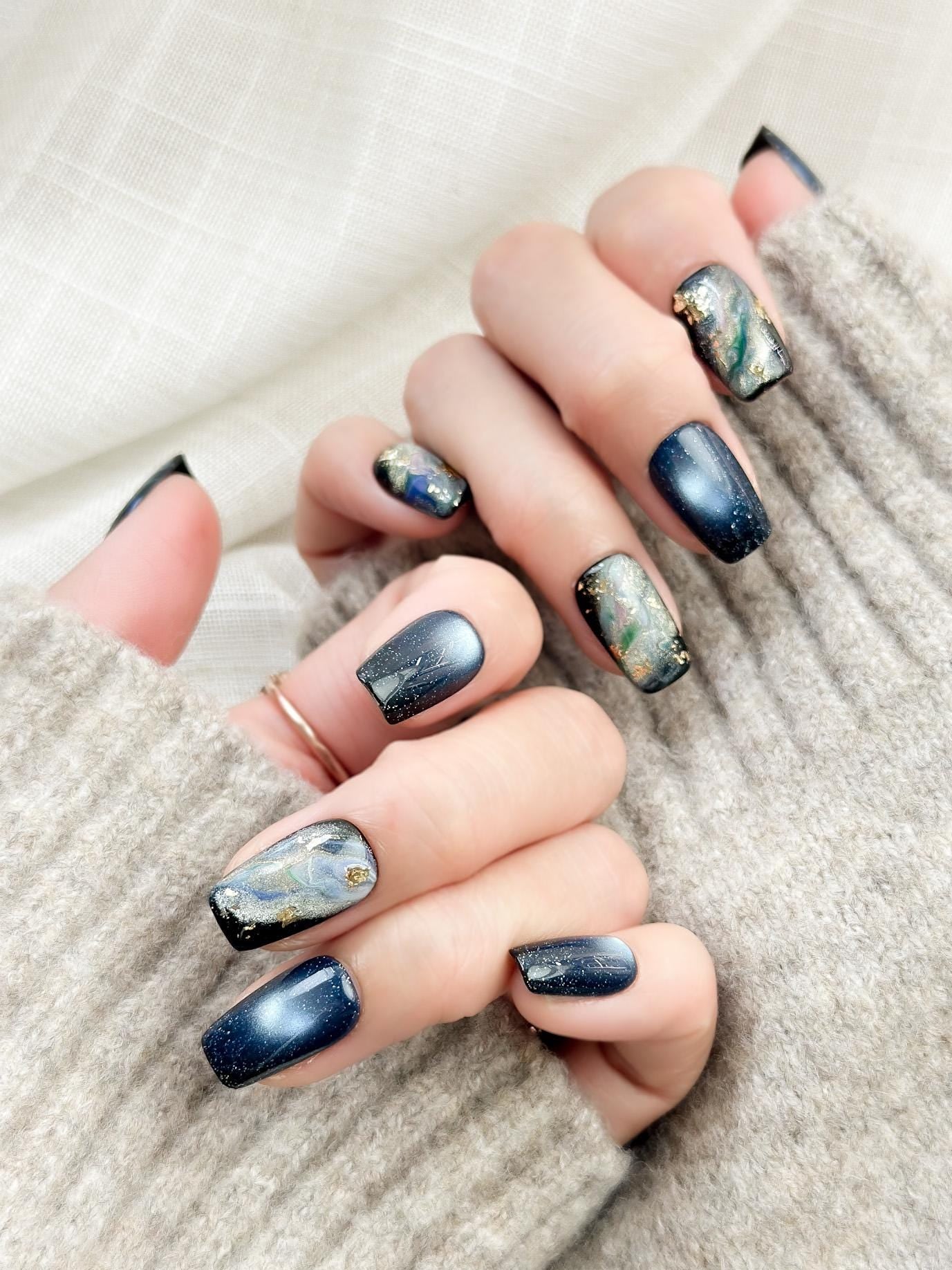 Hand with blue cat-eye press-on nails with galaxy shimmer and abstract marble accents in green, gold, and silver foil