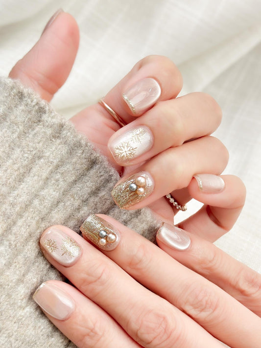 Hand with nude pink press-on nails with gold snowflake designs and glitter accents, featuring pearl embellishments for a festive holiday look