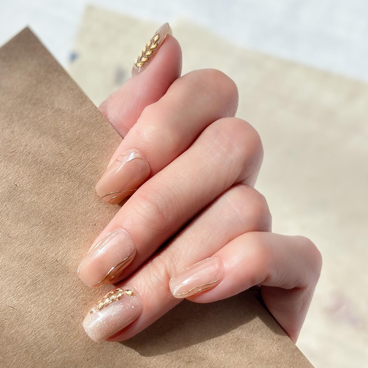 Hand with Nude beige press-on nails with gold leaf accents and glossy marble lines. Elegant neutral nail set with gold embellishments