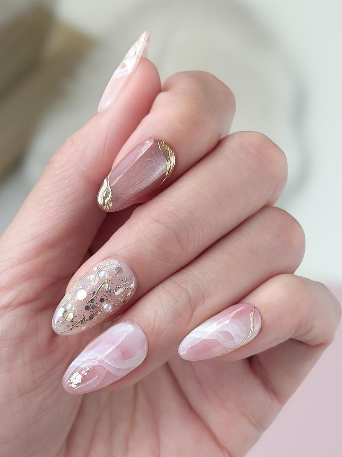 Close of of hand with Pink and nude luxury press-on nails with marble swirl designs, gold metallic accents, pearls, and glitter