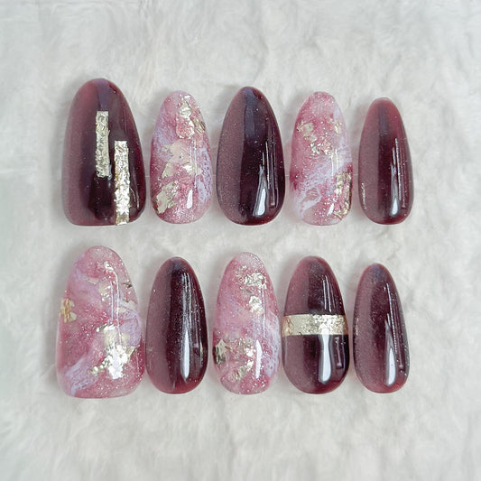 Almond-shaped press-on nails in deep plum and black cat-eye shimmer with pink marble accents, gold foil detailing, and glossy finish