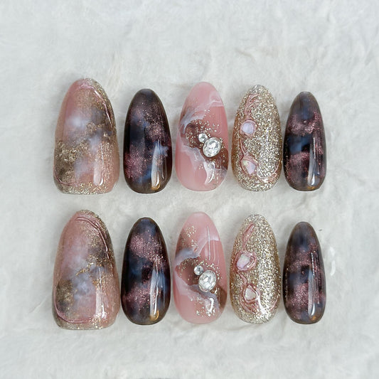 Almond-shaped luxury press-on nails in metallic brown and black cat-eye shimmer with pink marble accents, gold glitter, and pearl gemstone details