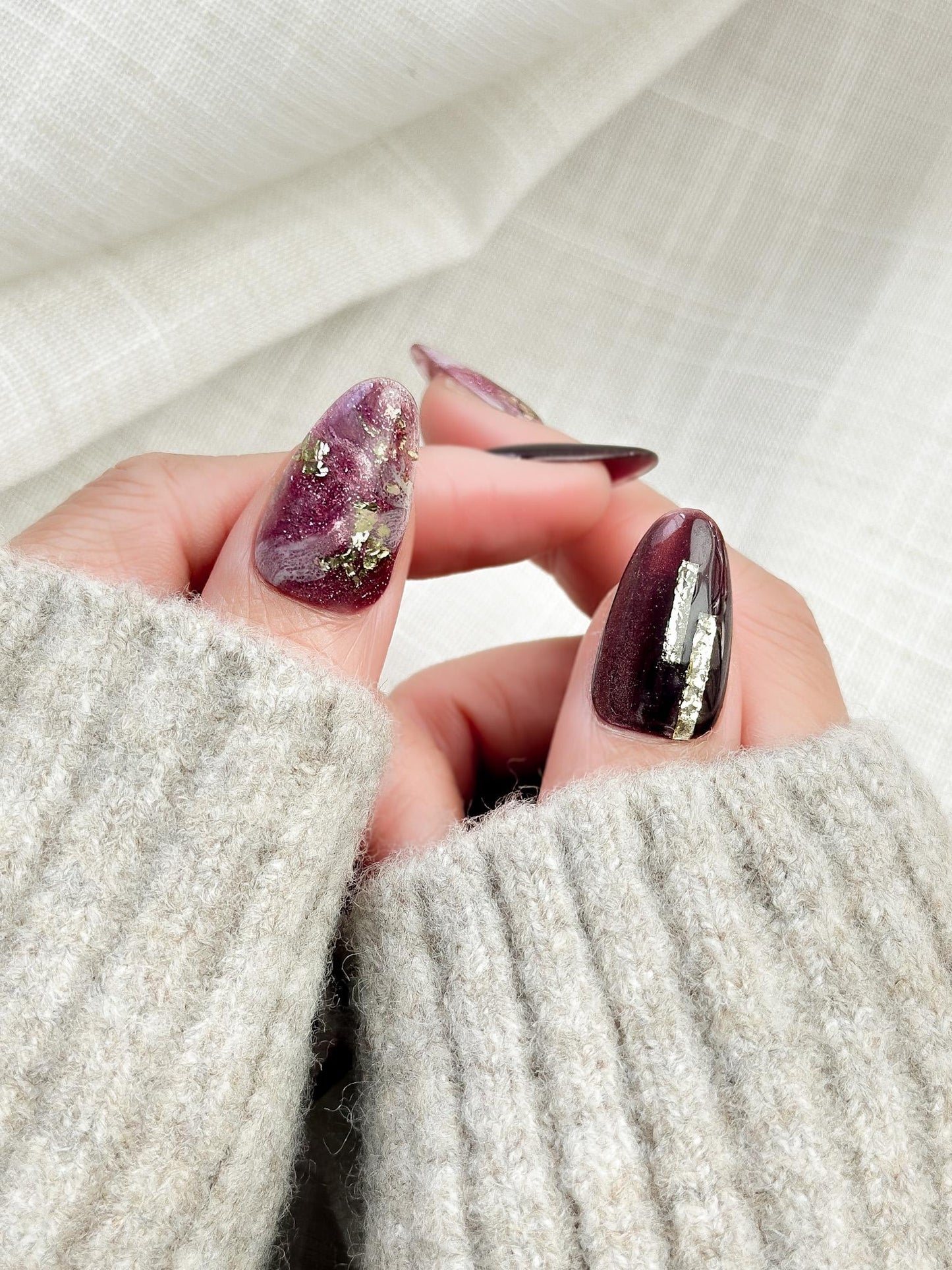 Hand with Almond-shaped press-on nails in deep plum cat-eye shimmer with pink and gold marble accents, gold foil detailing, and glossy finish