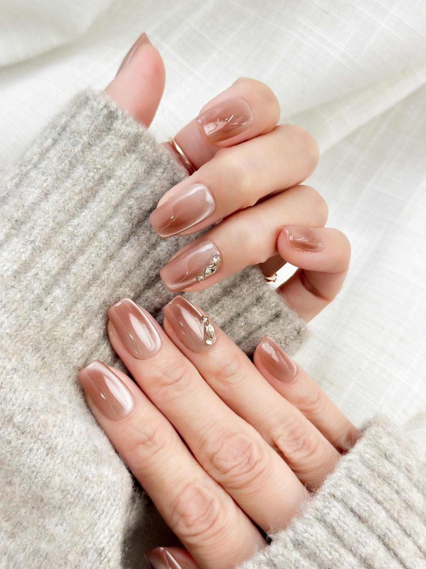 Close-up of a hand wearing glossy nude-brown press-on nails with a subtle cat-eye shimmer, featuring a gold leaf and crystal accent on one nail. The manicure is styled against a soft gray knit sweater for an elegant, cozy look