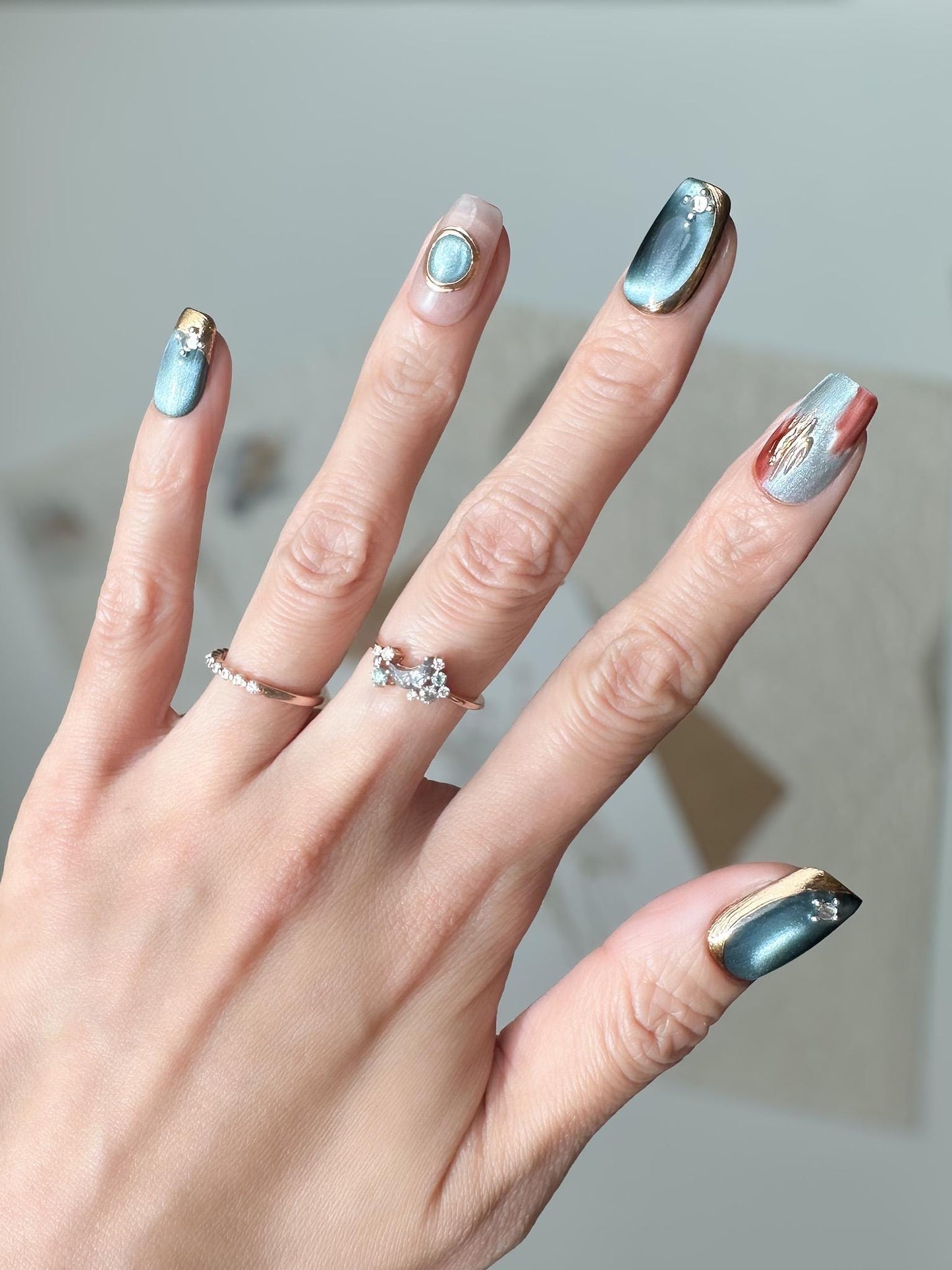 Close-up of hand with deep blue and silver chrome press-on nails with gold accents and gemstone details, featuring a metallic cat-eye shimmer