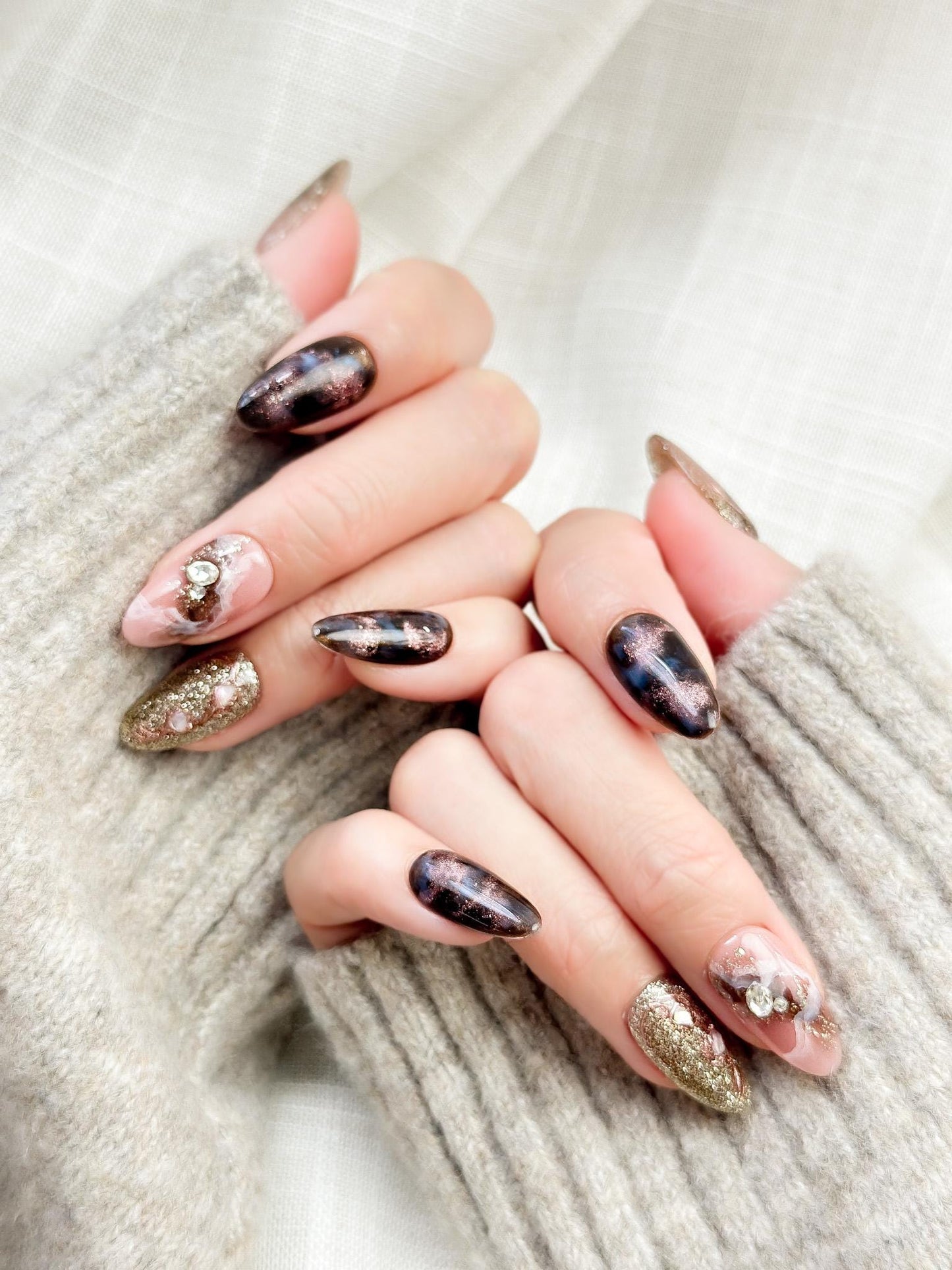 Hand with Almond-shaped luxury press-on nails in metallic brown and black cat-eye shimmer with pink marble accents, gold glitter, and pearl gemstone details