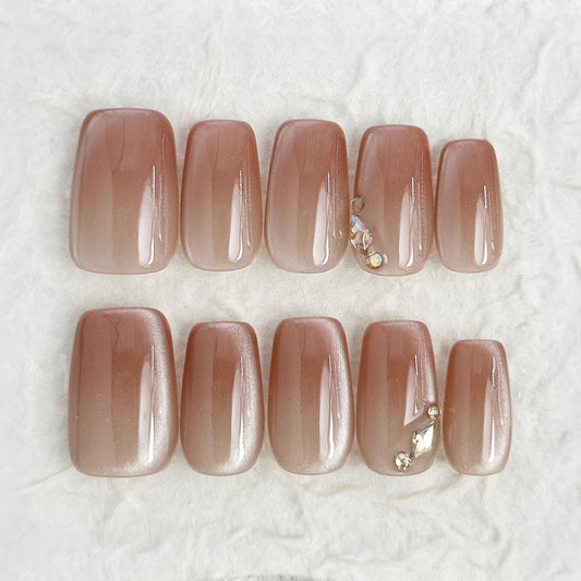 Close-up of a set of glossy nude-brown press-on nails with a subtle cat-eye shimmer, featuring a gold leaf and crystal accent on one nail.