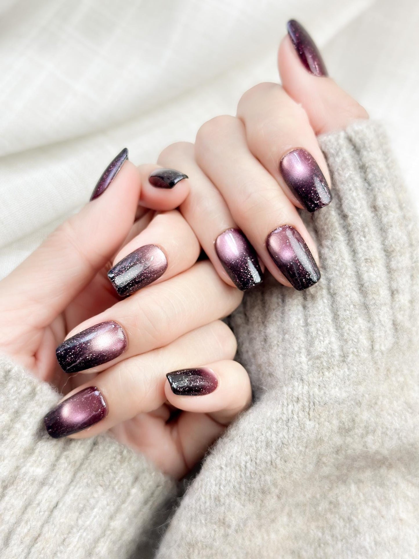 Hand with Short square press-on nails in a deep plum purple cat-eye shimmer with black gradient edges and silver sparkle accents