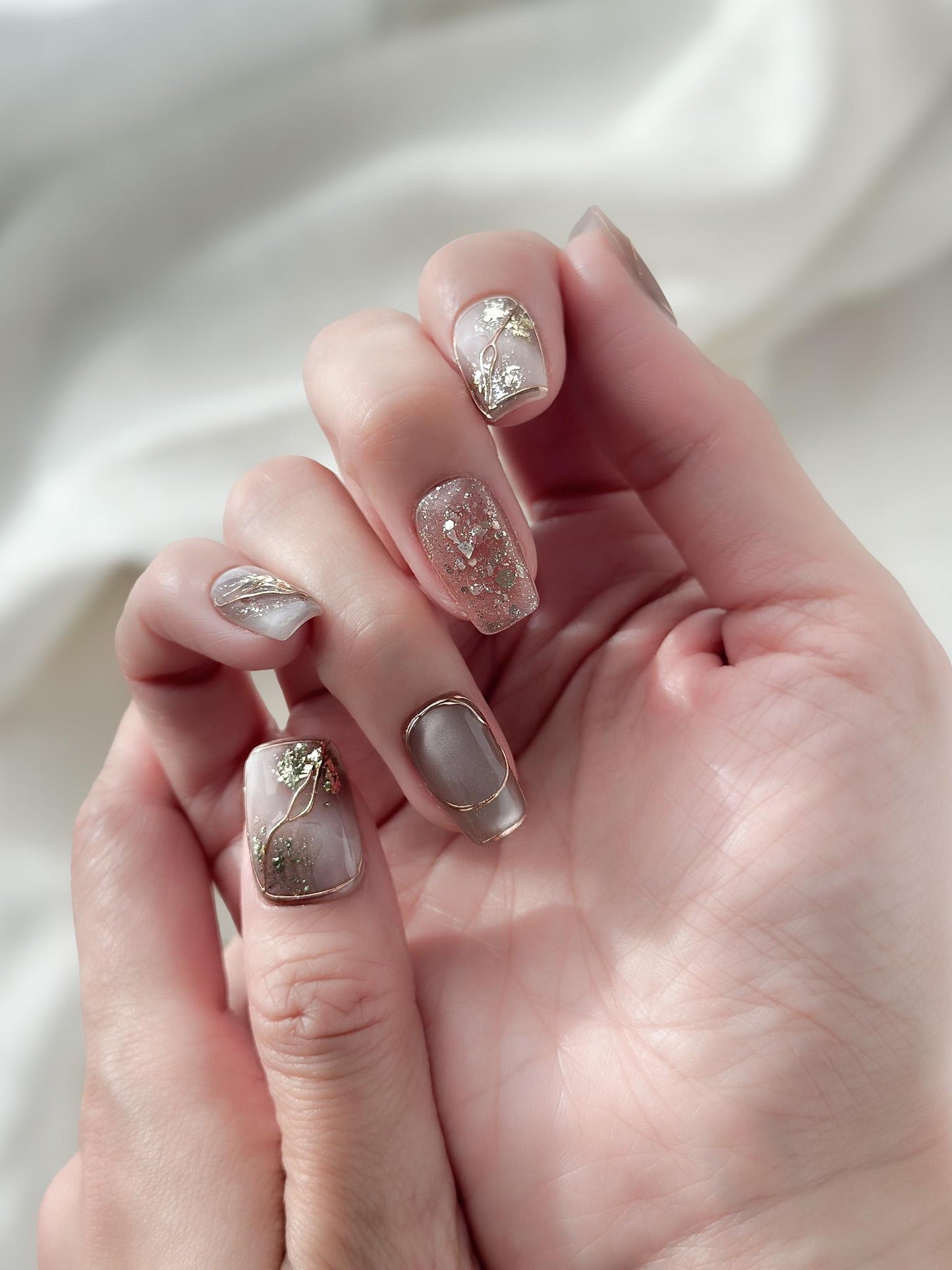 Close-up of hands with nude and taupe press-on nails with gold foil accents, rose gold glitter, and marble details