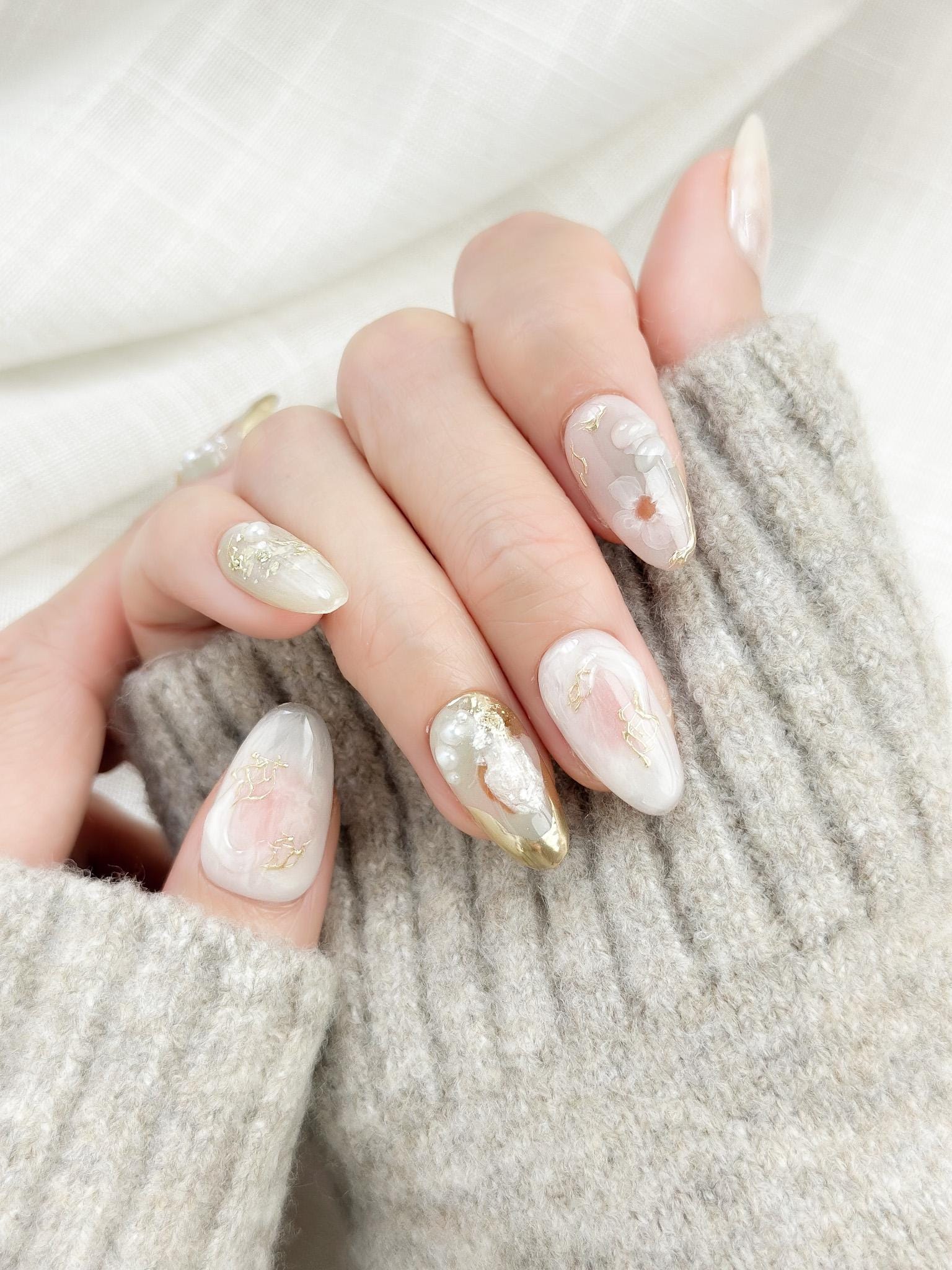 Hand with Almond-shaped white and pearlescent press-on nails with 3D floral accents, gold chrome details, pearl embellishments, and soft milky marble art