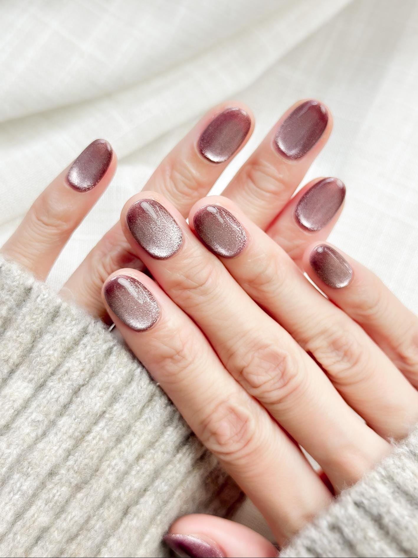 Short oval press-on nails in a metallic mauve cat-eye shimmer, glossy finish, luxury reusable press-on manicure by ManiFleur. Elegant mauve shimmer nails styled against a cozy knit sweater
