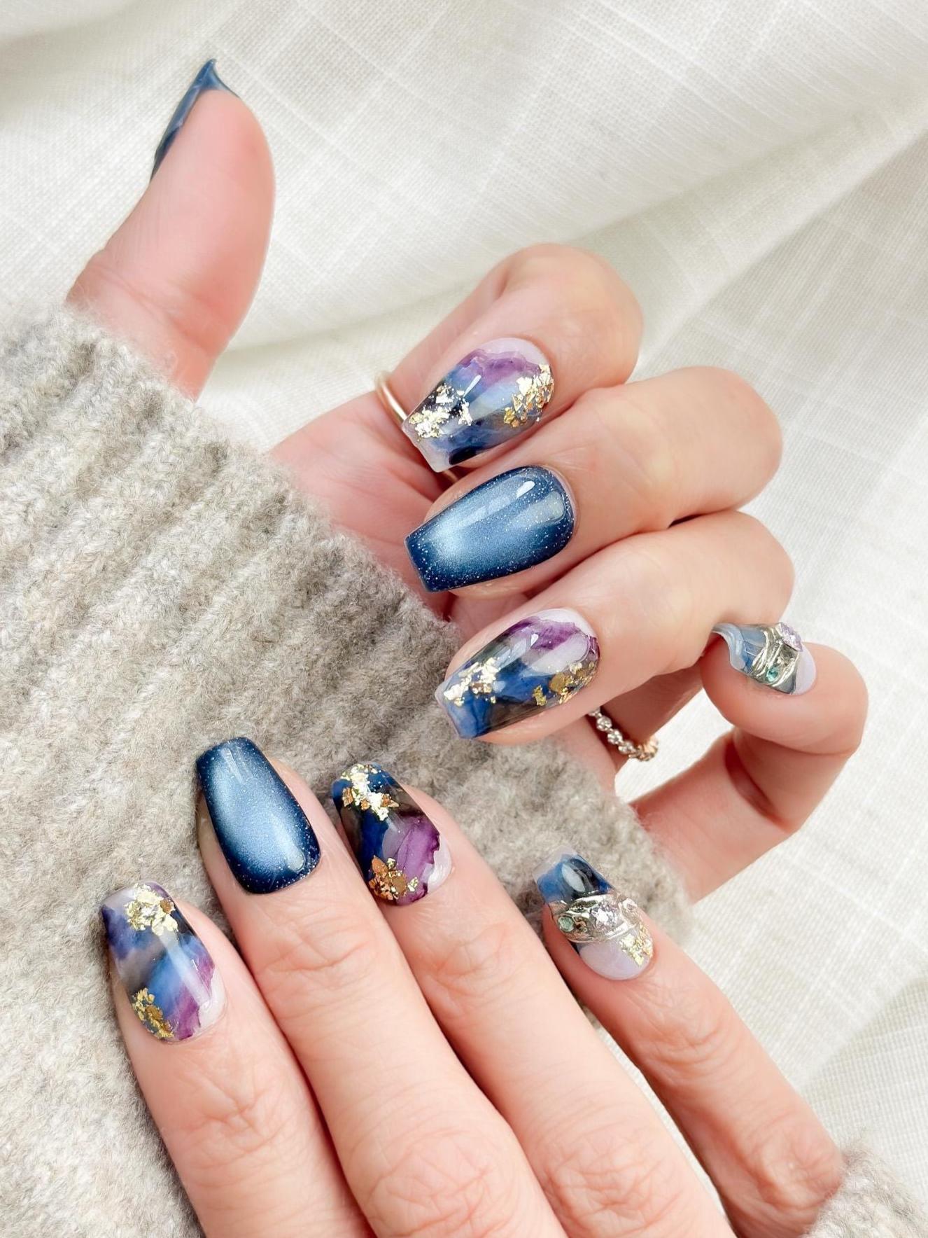 Hand with Blue galaxy press-on nails with starry cat-eye shimmer and watercolor marble accents in navy, lavender, and white, finished with gold foil details