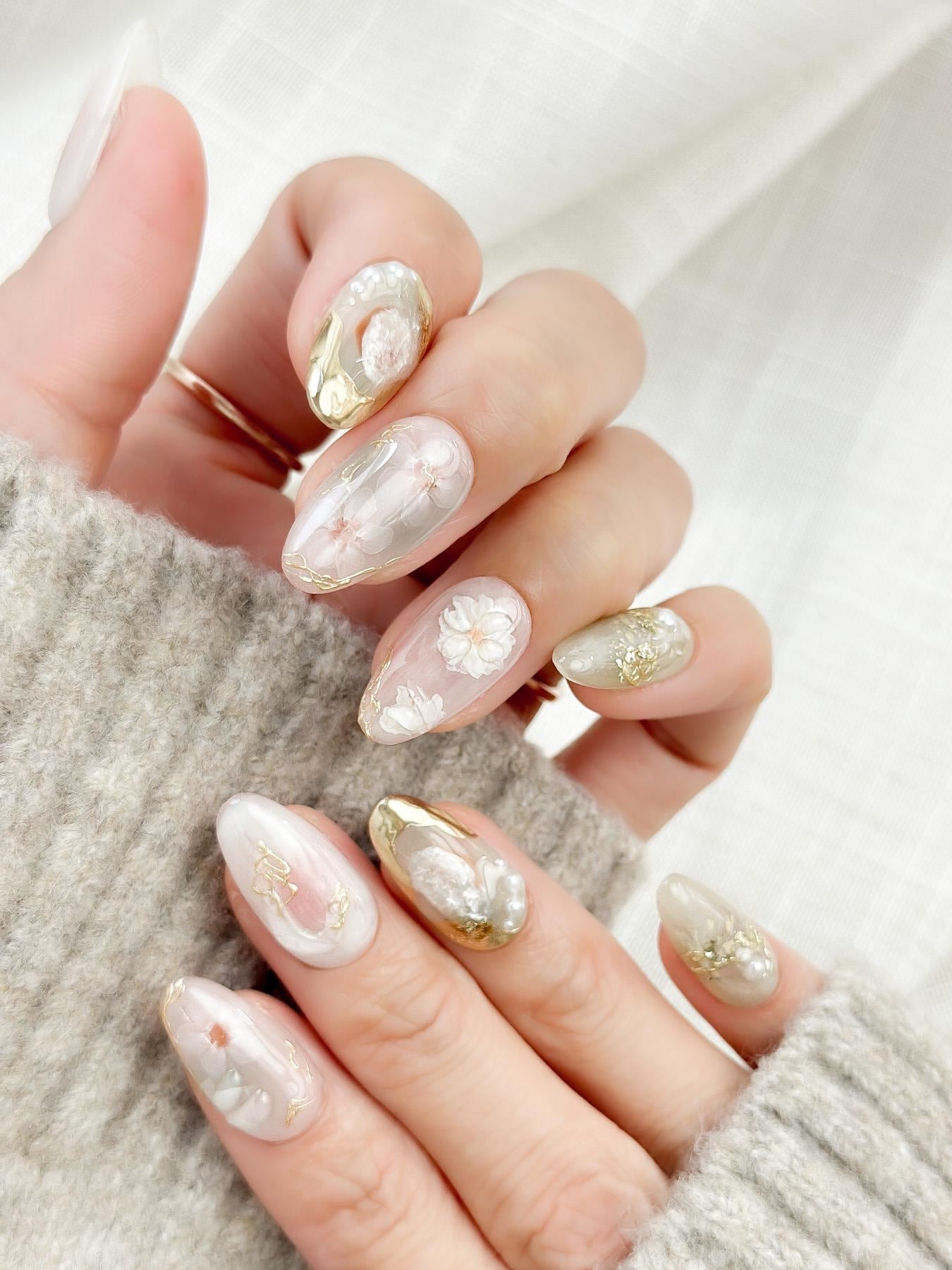 Hand with Almond-shaped white and pearlescent press-on nails with 3D floral accents, gold chrome details, pearl embellishments, and soft milky marble art
