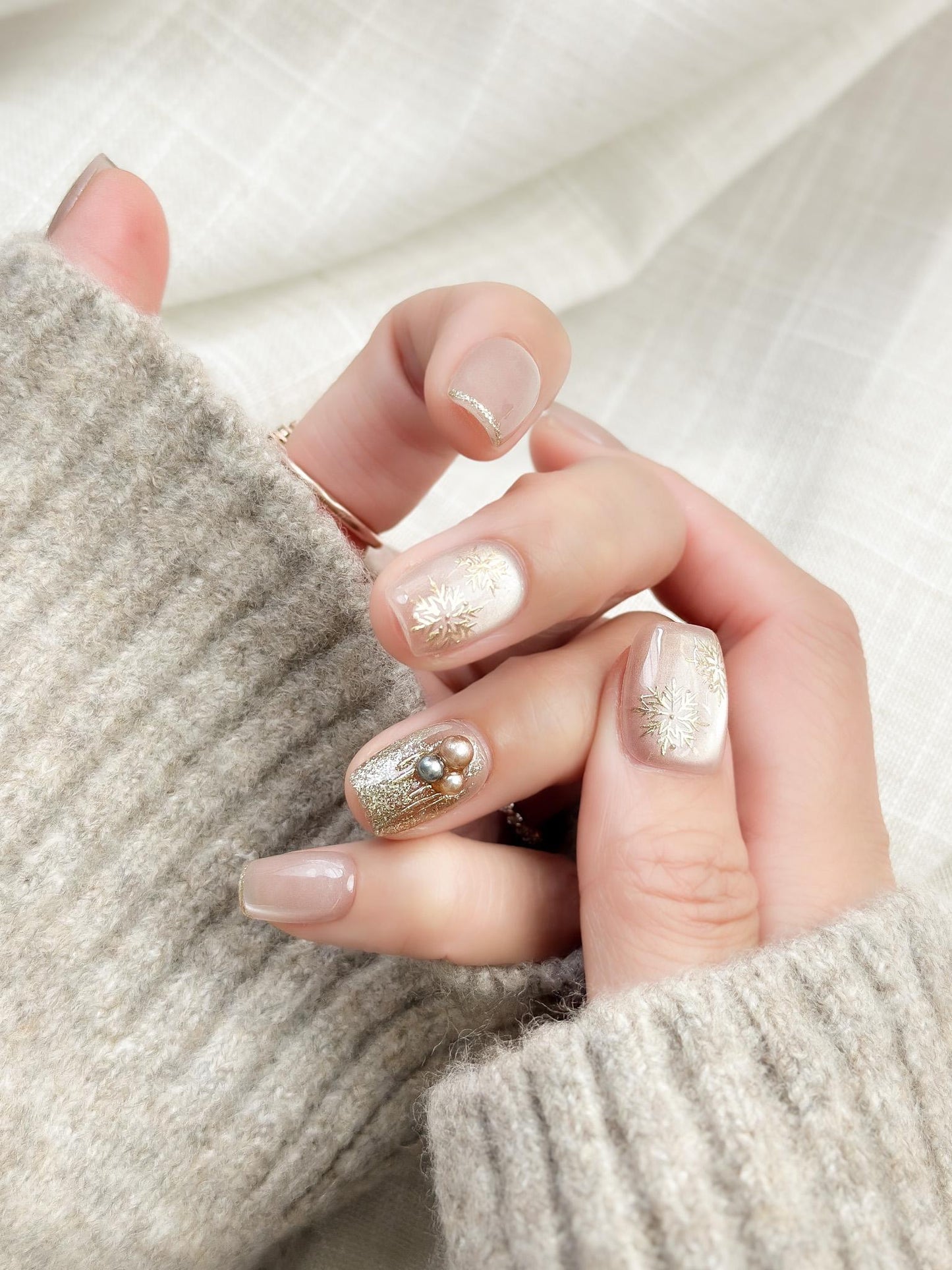 Hand with nude pink press-on nails with gold snowflake designs and glitter accents, featuring pearl embellishments for a festive holiday look