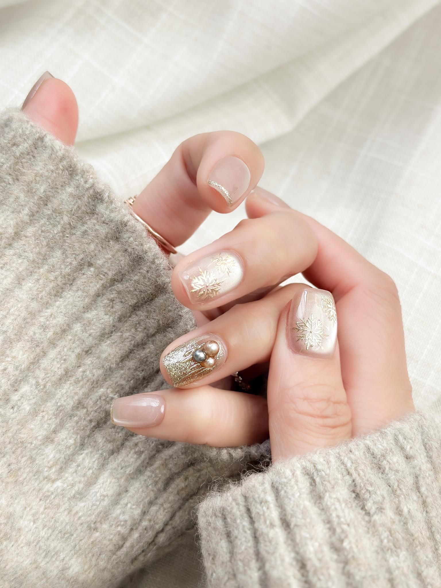 Hand with nude pink press-on nails with gold snowflake designs and glitter accents, featuring pearl embellishments for a festive holiday look