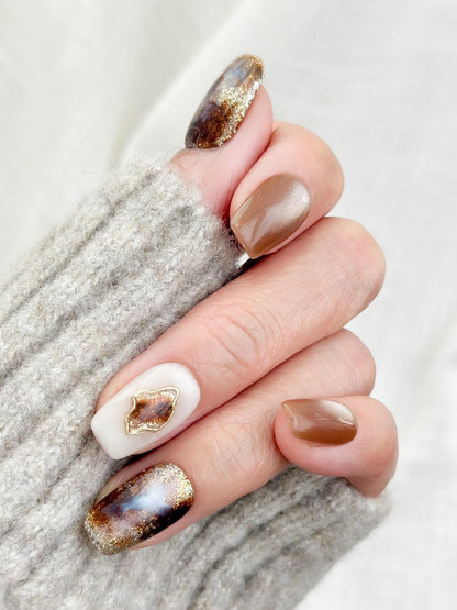 Hand with brown and ivory press-on nails in tortoise pattern, with gold foil detailing, and glossy cat-eye shimmer