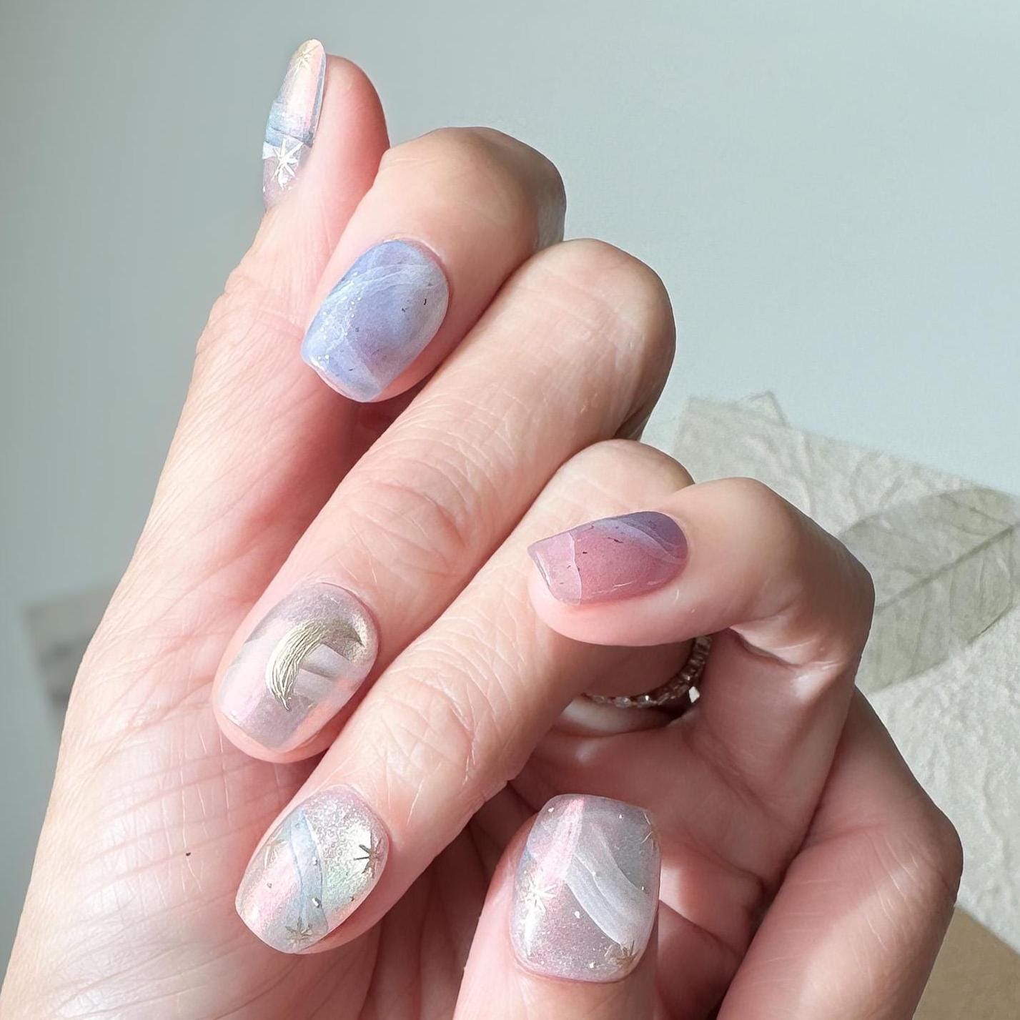 Hand with Pastel celestial press-on nails with pink, lavender, and blue marble swirls, gold star accents, and a gold crescent moon design