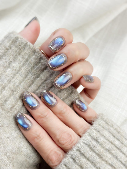Hand with Square shaped grey-blue cat eye press-on nails with metallic shimmer and gold foil border accents