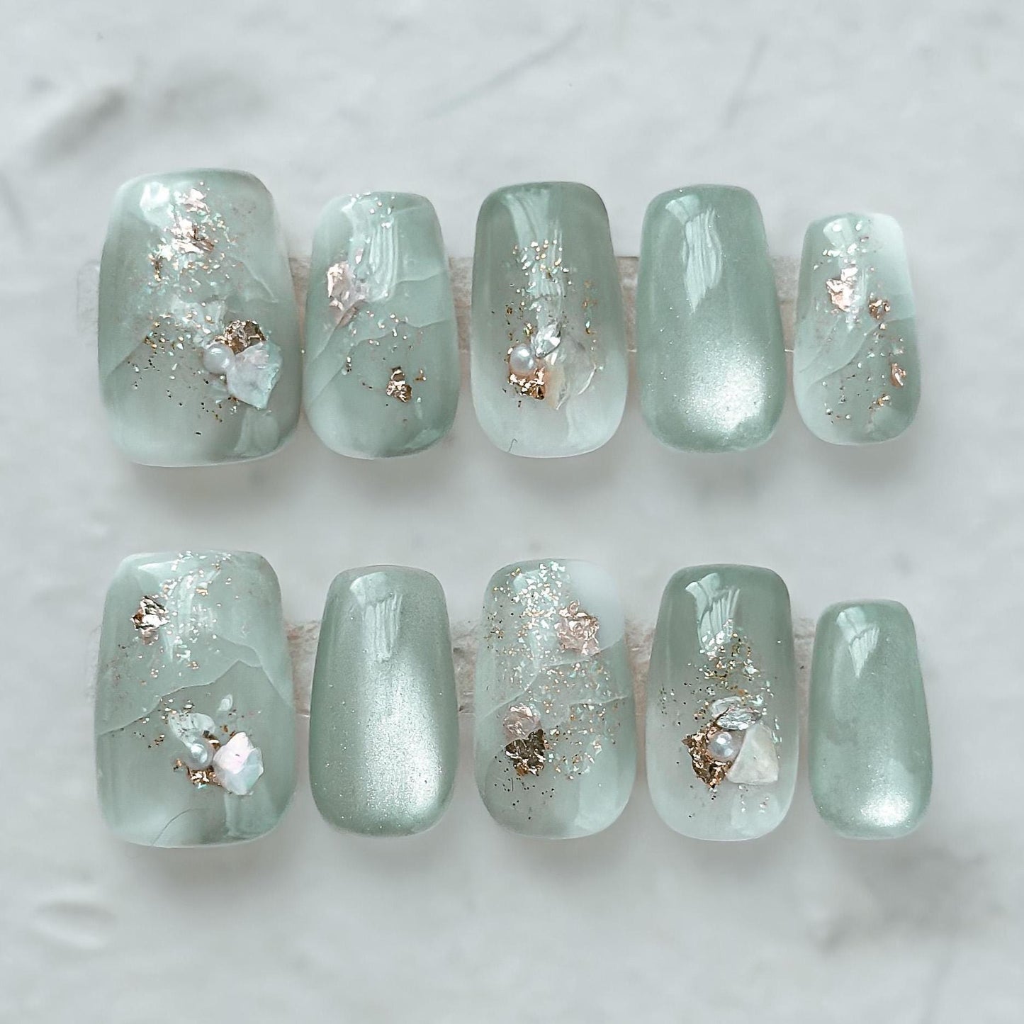 Mint green press-on nails with pearlescent shimmer, marble accents, gold foil, and tiny pearl embellishments