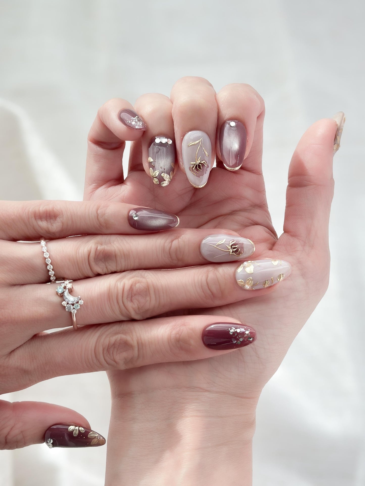 Hand with almond shaped Press-on nails in deep plum and mauve tones with gold leaf accents, metallic floral designs, and silver gemstone details