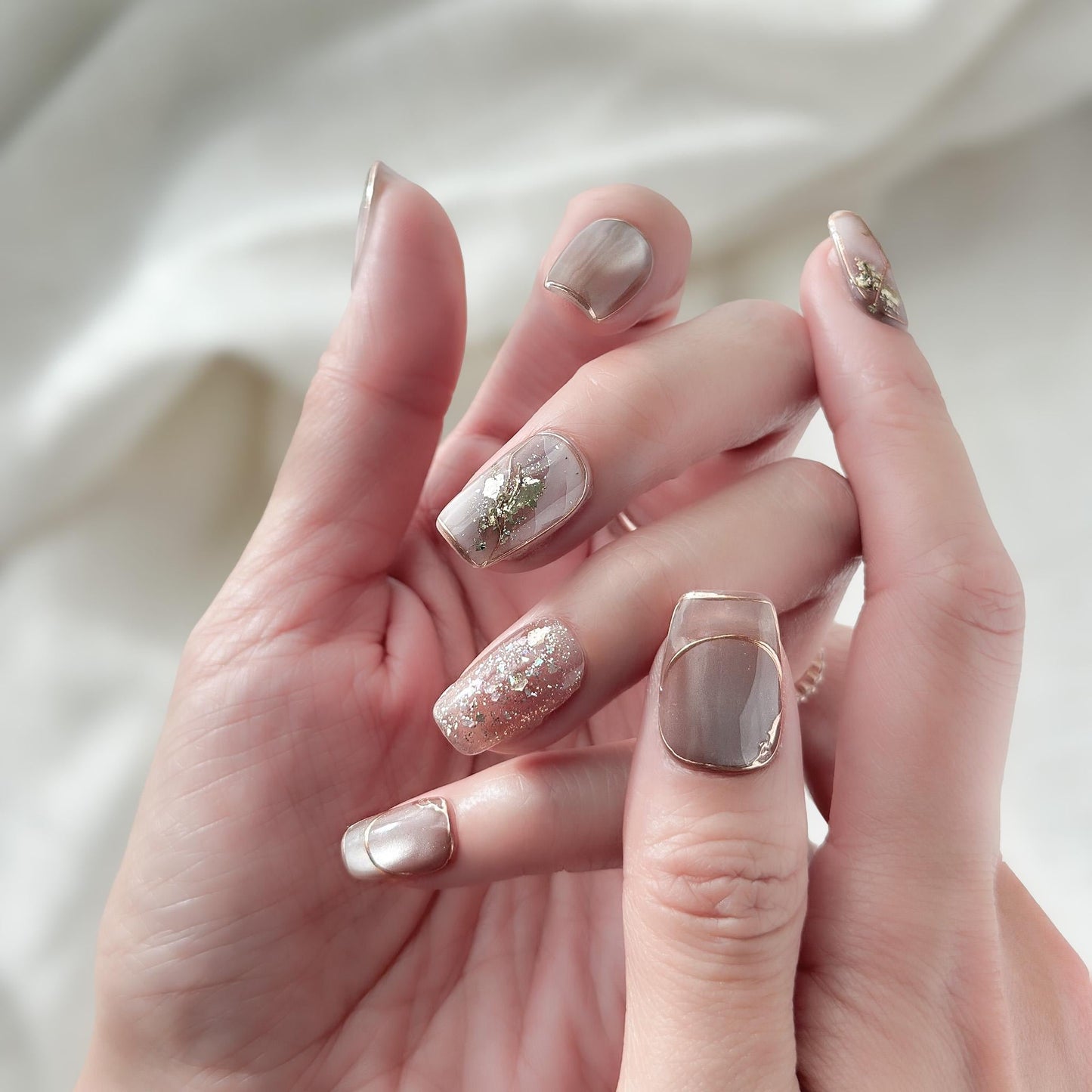 Close-up of hands with nude and taupe press-on nails with gold foil accents, rose gold glitter, and marble details