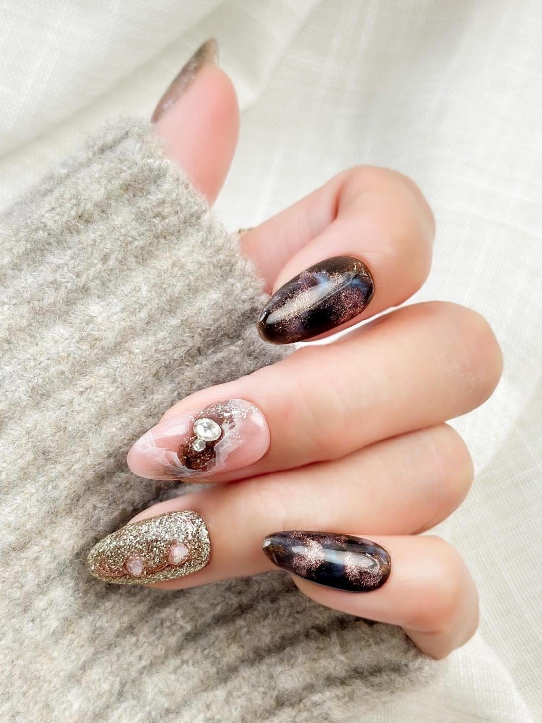 Hand with Almond-shaped luxury press-on nails in metallic brown and black cat-eye shimmer with pink marble accents, gold glitter, and pearl gemstone details