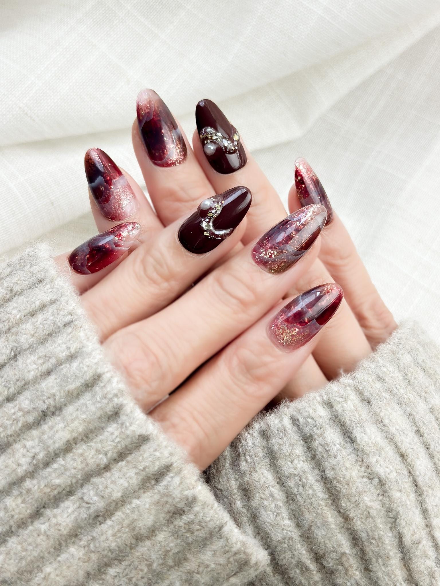 Hand with Almond-shaped burgundy and wine-red press-on nails with glossy finish, abstract marble art, gold glitter accents, and crystal pearl embellishments.
