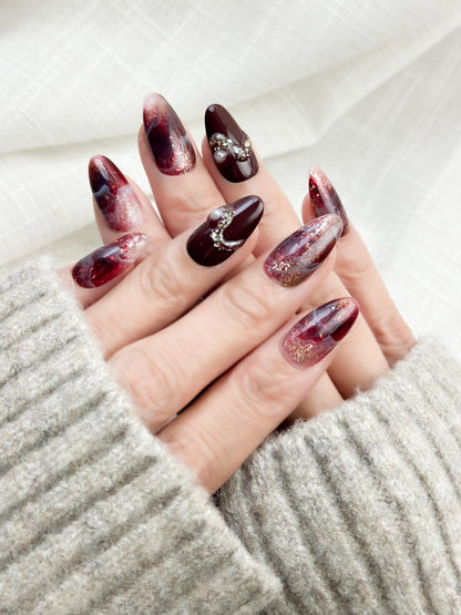 Hand with Almond-shaped burgundy and wine-red press-on nails with glossy finish, abstract marble art, gold glitter accents, and crystal pearl embellishments.
