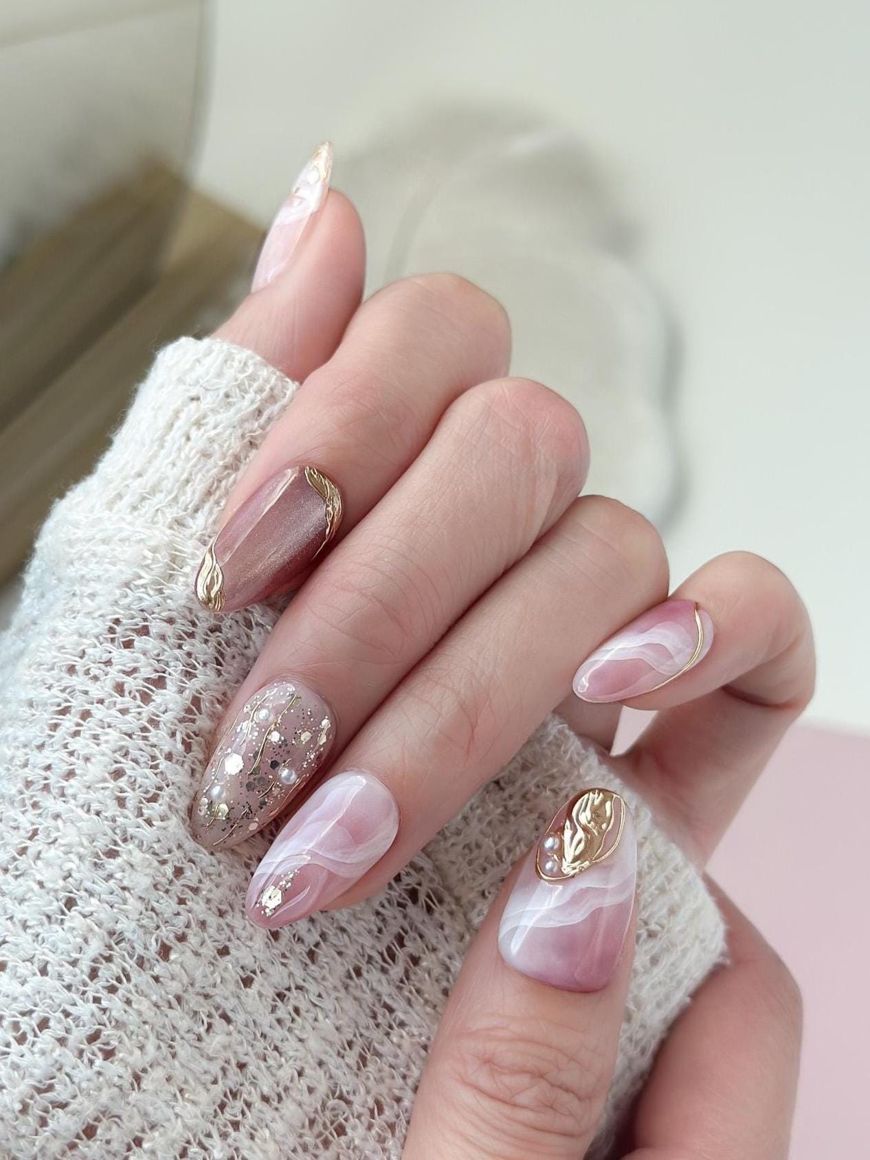 Close of of hand with Pink and nude luxury press-on nails with marble swirl designs, gold metallic accents, pearls, and glitter