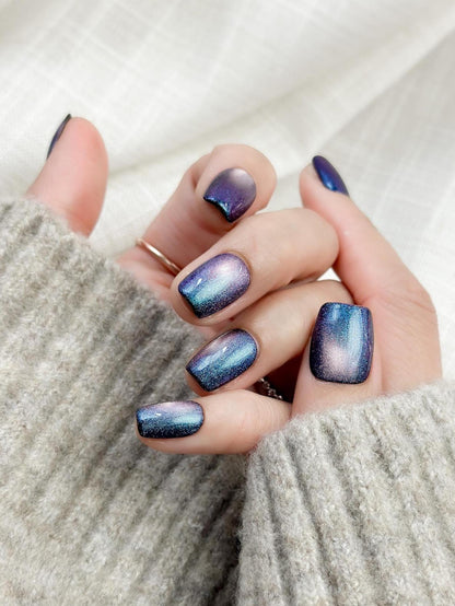 Hand with Short square shaped cat-eye press-on nails in shifting blue, purple, and teal galaxy shimmer
