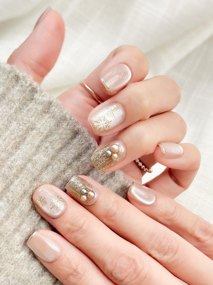 Hand with nude pink press-on nails with gold snowflake designs and glitter accents, featuring pearl embellishments for a festive holiday look