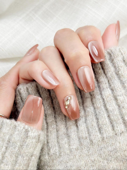 Close-up of a hand wearing glossy nude-brown press-on nails with a subtle cat-eye shimmer, featuring a gold leaf and crystal accent on one nail. The manicure is styled against a soft gray knit sweater for an elegant, cozy look
