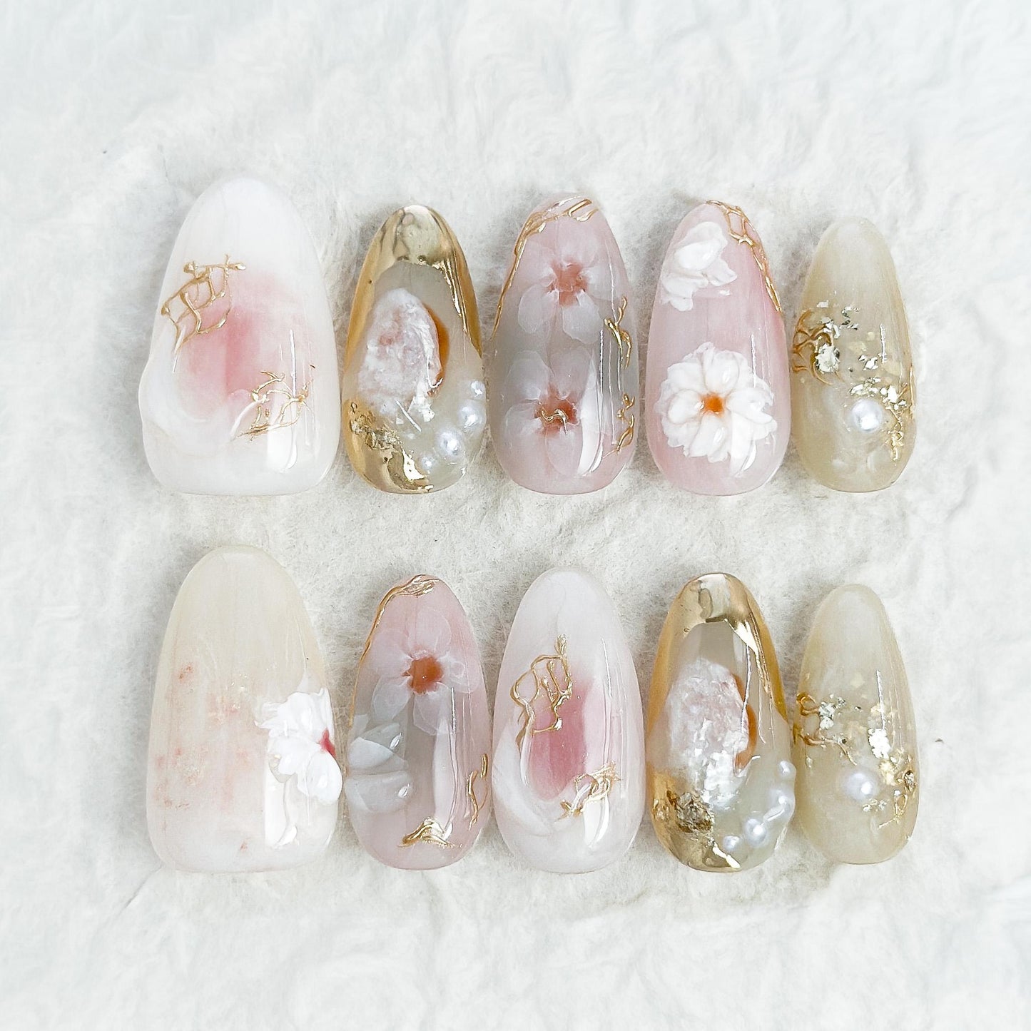 Almond-shaped white and pearlescent press-on nails with 3D floral accents, gold chrome details, pearl embellishments, and soft milky marble art