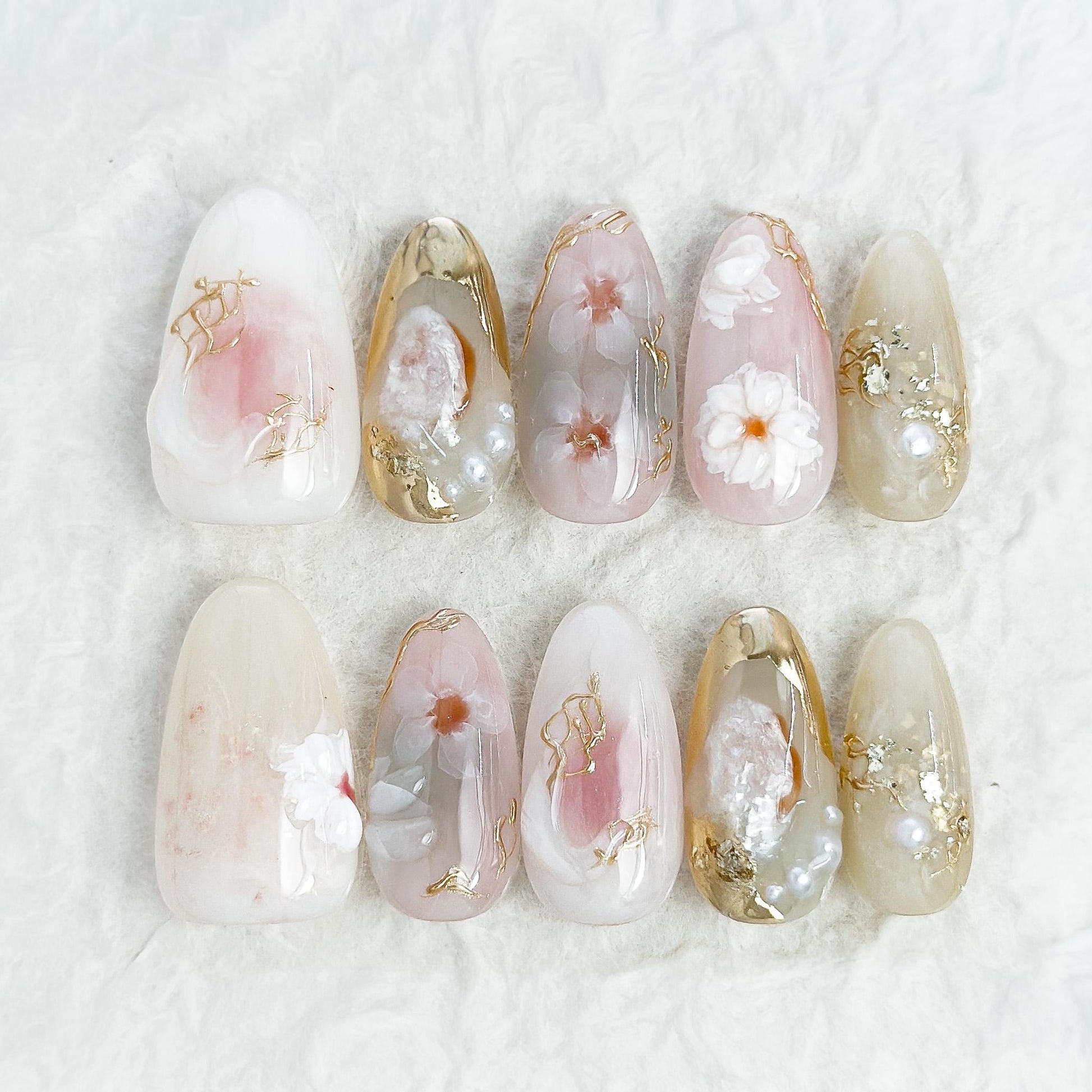 Almond-shaped white and pearlescent press-on nails with 3D floral accents, gold chrome details, pearl embellishments, and soft milky marble art