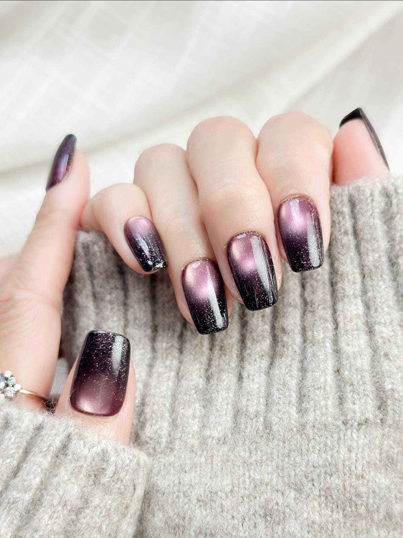 Hand with Short square press-on nails in a deep plum purple cat-eye shimmer with black gradient edges and silver sparkle accents