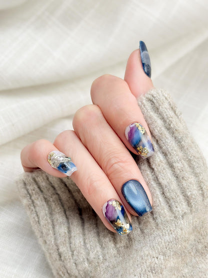 Hand with Blue galaxy press-on nails with starry cat-eye shimmer and watercolor marble accents in navy, lavender, and white, finished with gold foil details