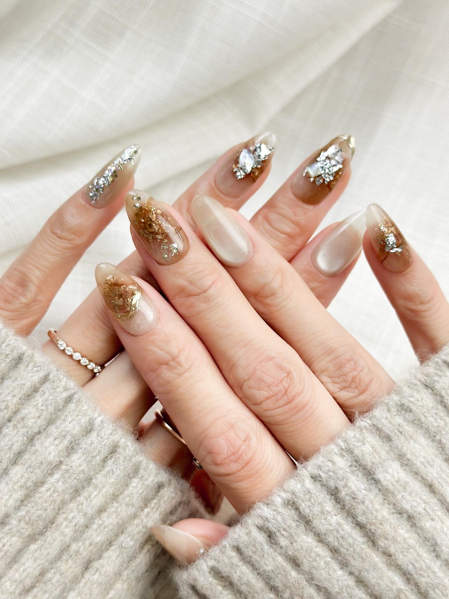 Close-up of almond-shaped nude and caramel brown press-on nails with gold foil accents and subtle shimmer, styled against a soft beige knit sweater