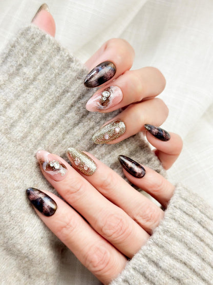 Hand with Almond-shaped luxury press-on nails in metallic brown and black cat-eye shimmer with pink marble accents, gold glitter, and pearl gemstone details