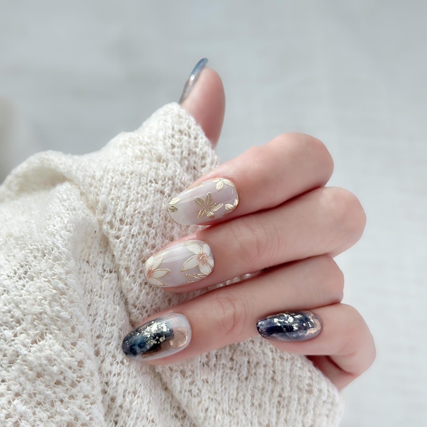 Hand with Oval-shaped press-on nails with deep navy and gold marble art, gold foil accents, and soft floral details