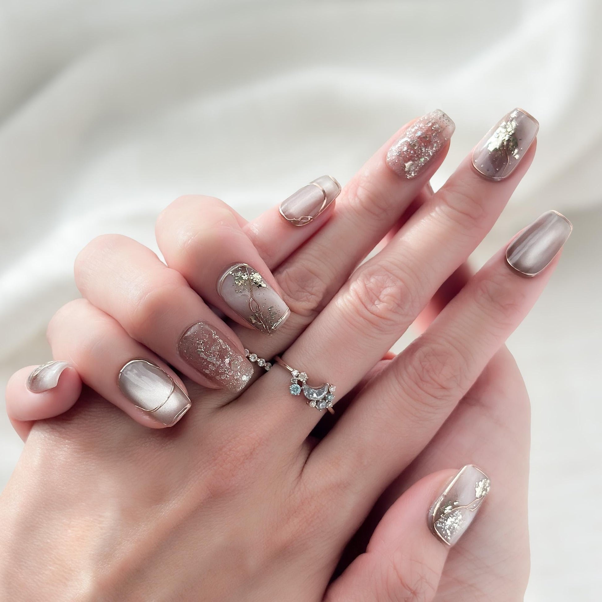 Close-up of hands with nude and taupe press-on nails with gold foil accents, rose gold glitter, and marble details
