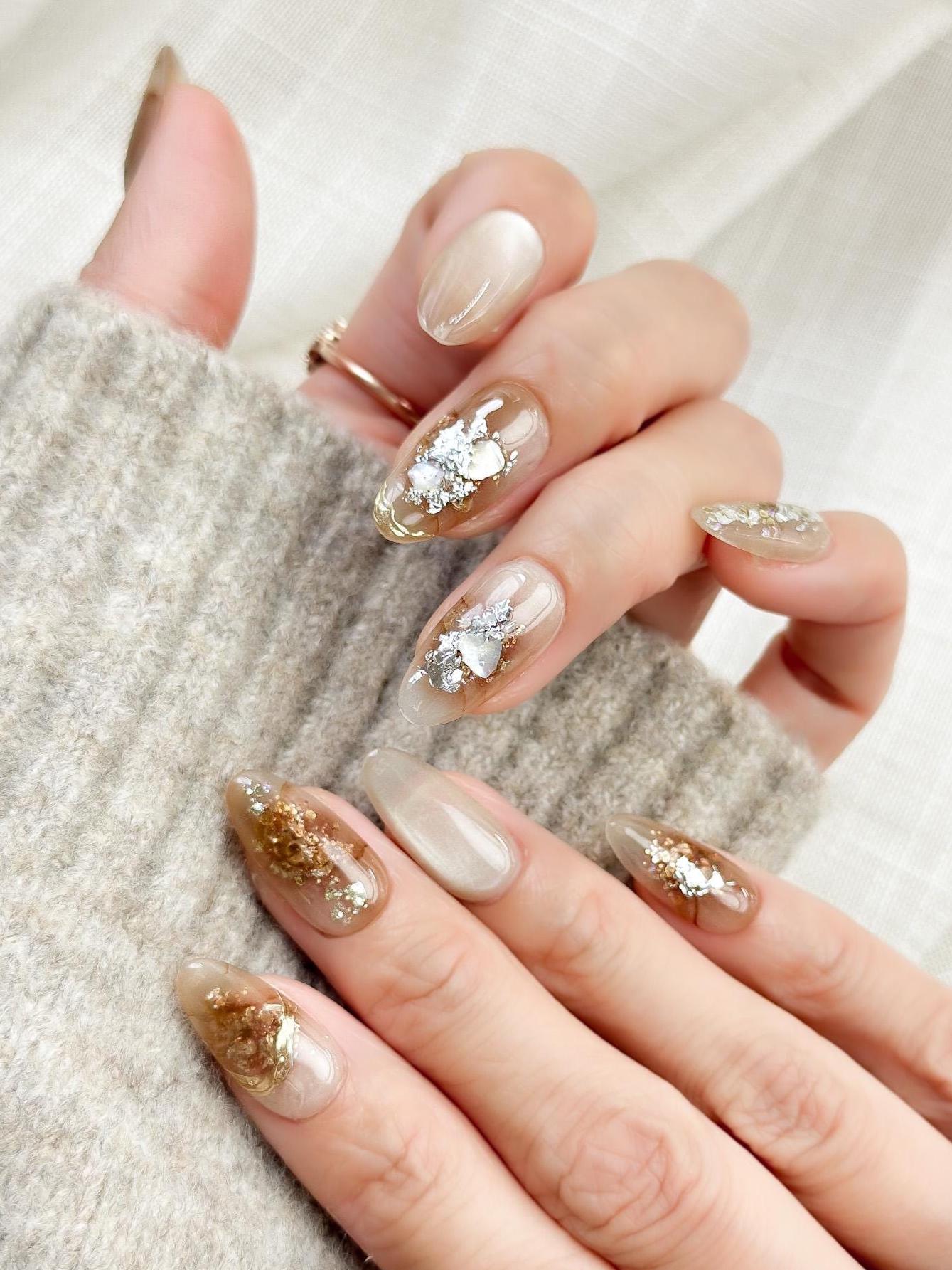 Close-up of almond-shaped nude and caramel brown press-on nails with gold foil accents and subtle shimmer, styled against a soft beige knit sweater