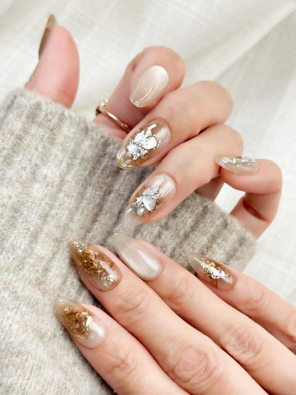 Close-up of almond-shaped nude and caramel brown press-on nails with gold foil accents and subtle shimmer, styled against a soft beige knit sweater