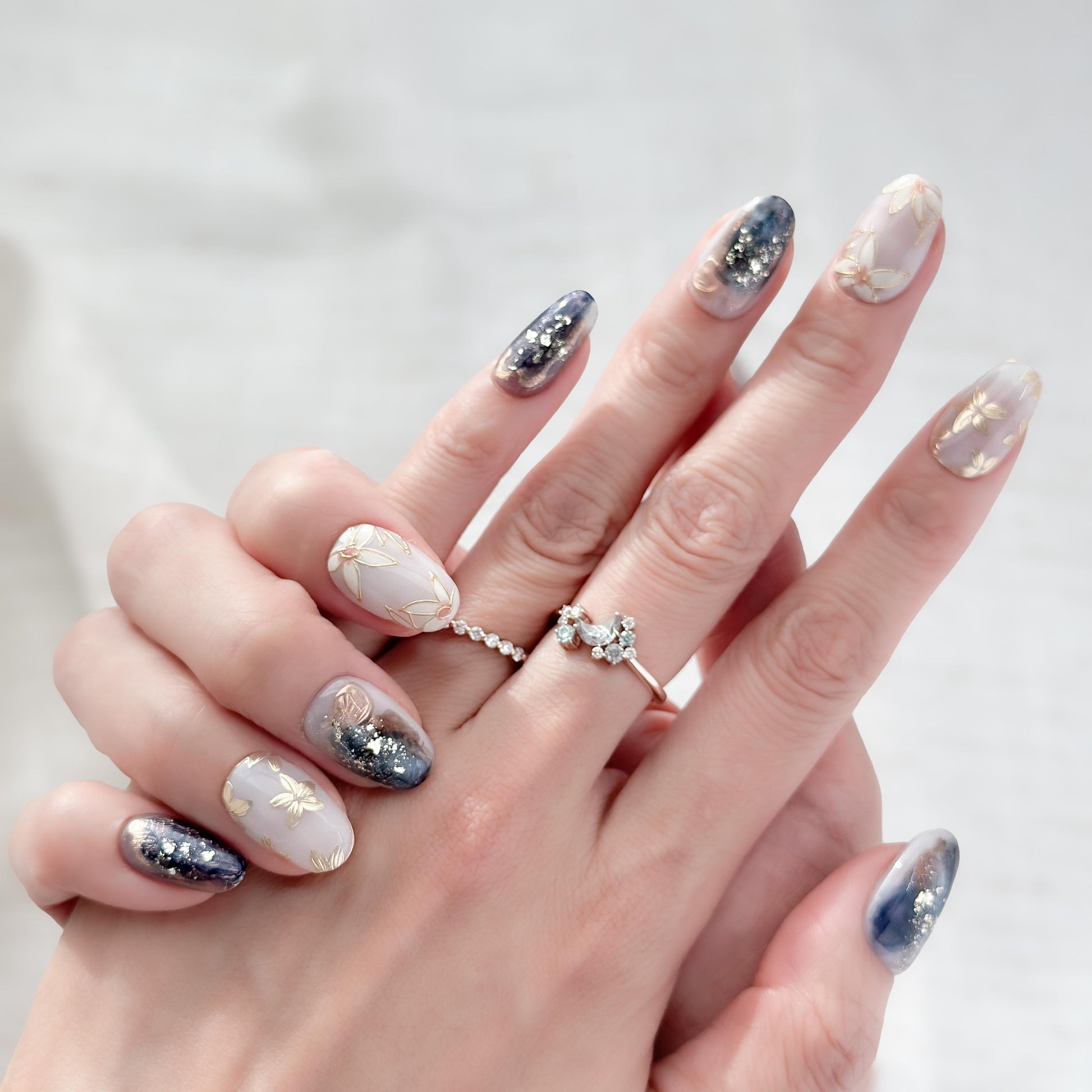 Hand with Oval-shaped press-on nails with deep navy and gold marble art, gold foil accents, and soft floral details.