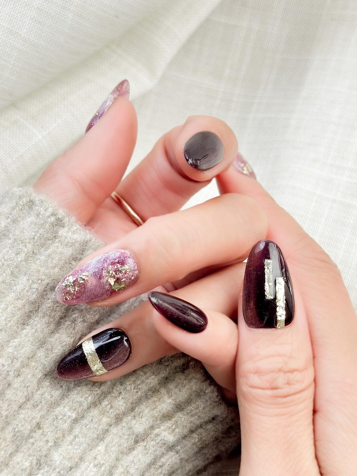 Hand with Almond-shaped press-on nails in deep plum cat-eye shimmer with pink and gold marble accents, gold foil detailing, and glossy finish