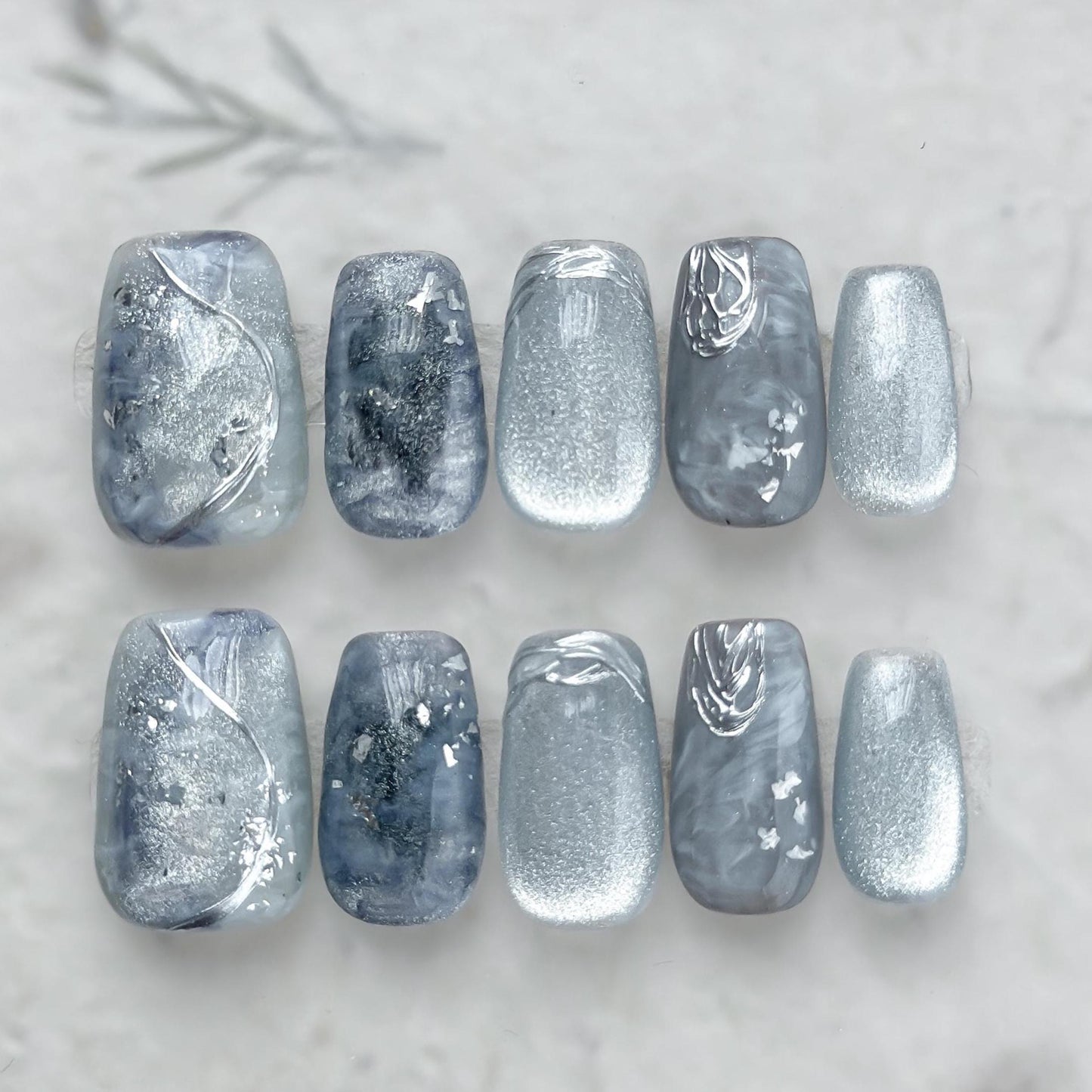 Blue and silver marble press-on nails with icy shimmer, metallic accents, and scattered silver foil flakes. Winter-inspired design featuring frosted blue hues and glossy snow-like details