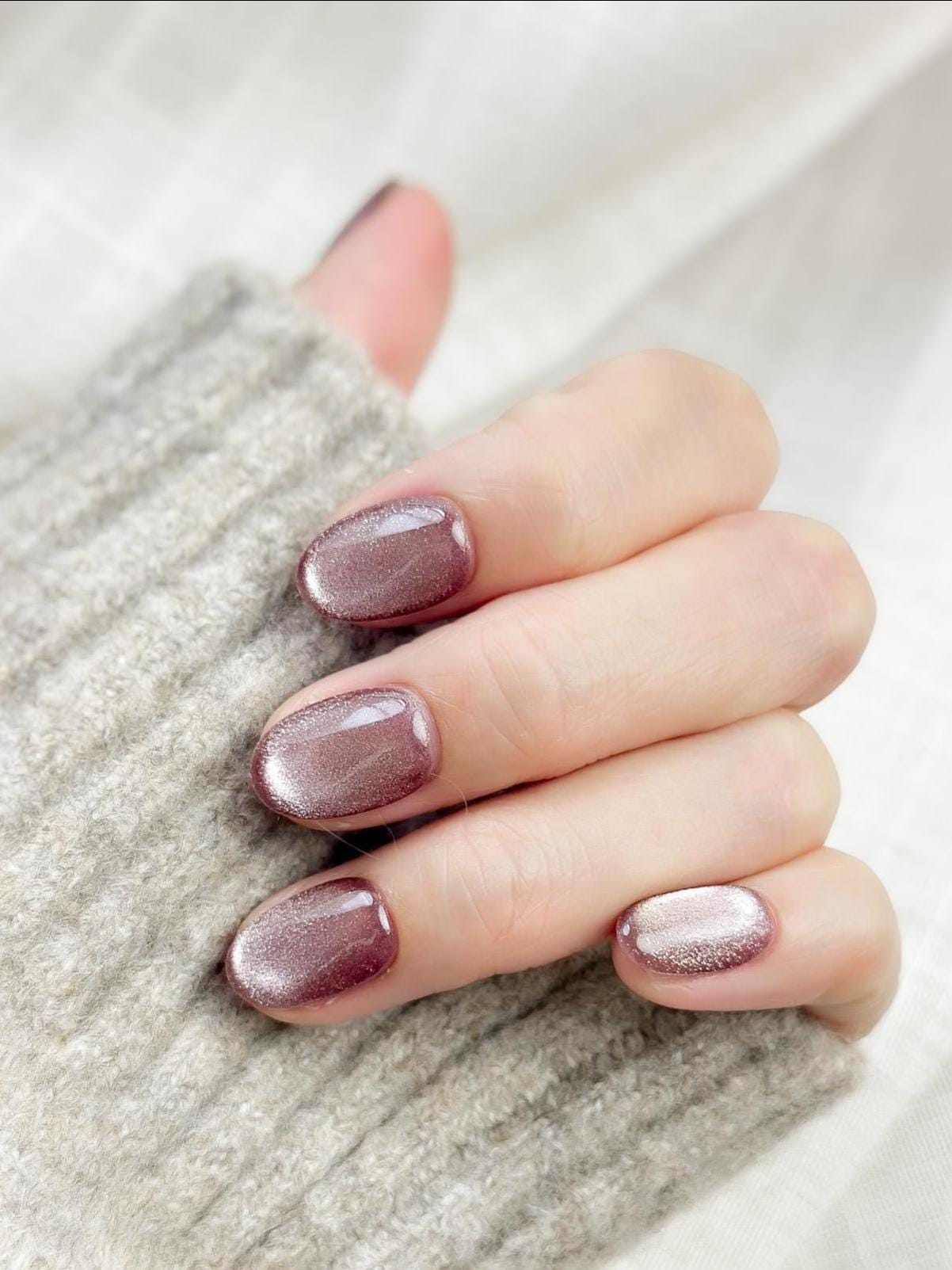 Short oval press-on nails in a metallic mauve cat-eye shimmer, glossy finish, luxury reusable press-on manicure by ManiFleur. Elegant mauve shimmer nails styled against a cozy knit sweater