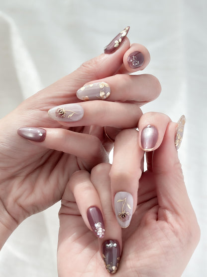 Hand with almond shaped Press-on nails in deep plum and mauve tones with gold leaf accents, metallic floral designs, and silver gemstone details