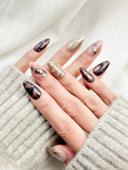 Hand with Almond-shaped luxury press-on nails in metallic brown and black cat-eye shimmer with pink marble accents, gold glitter, and pearl gemstone details