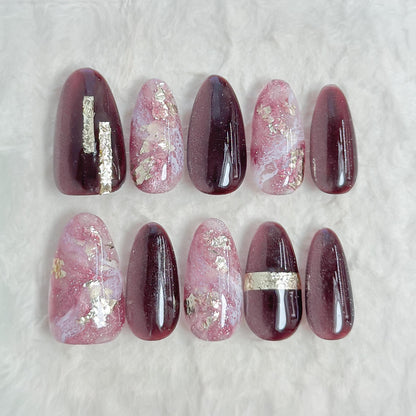 Almond-shaped press-on nails in deep plum and black cat-eye shimmer with pink marble accents, gold foil detailing, and glossy finish